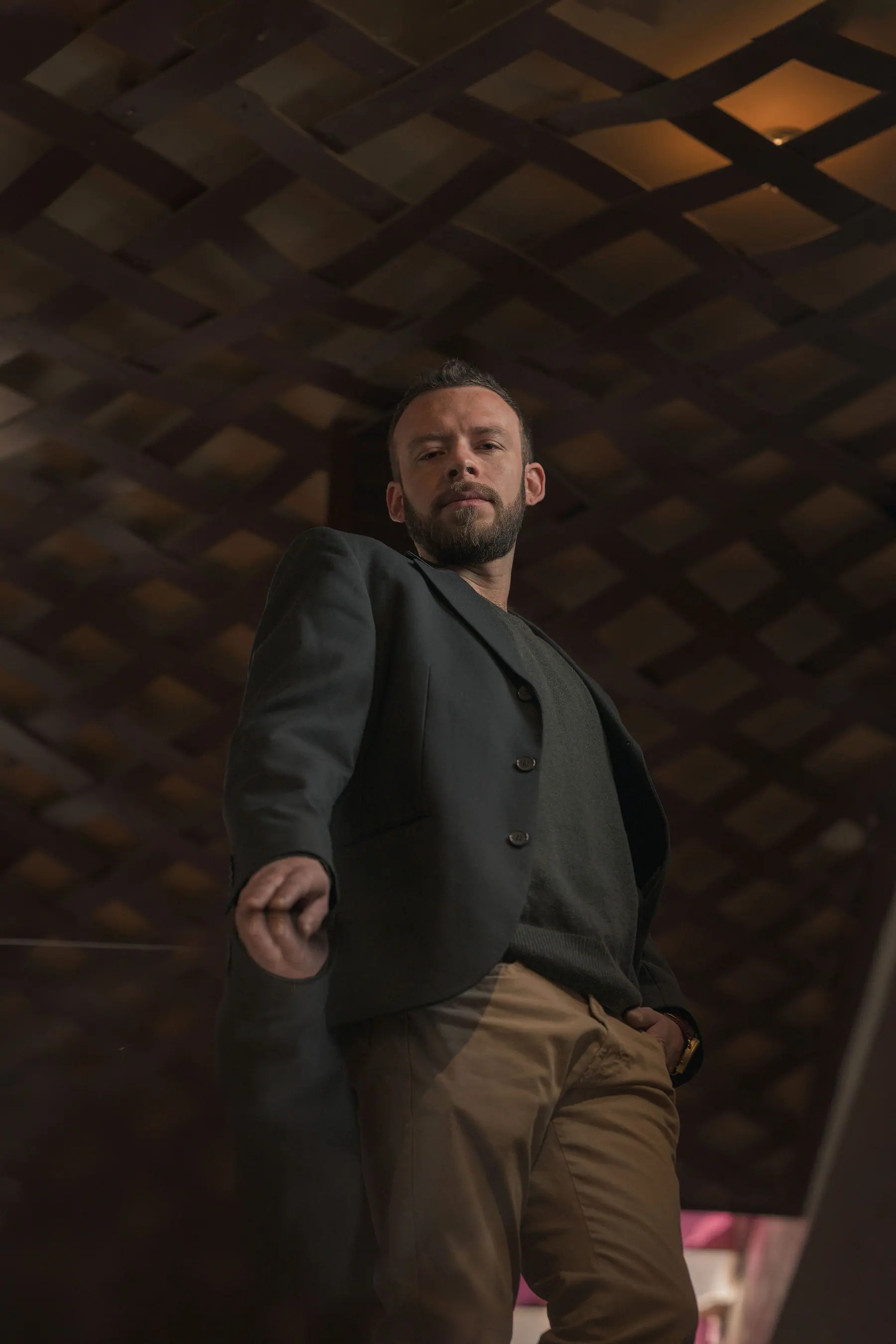 Elex Asbun avanzando hacia la cámara con blazer verde oscuro y pantalón camel, ante una celosía de madera en penumbra.