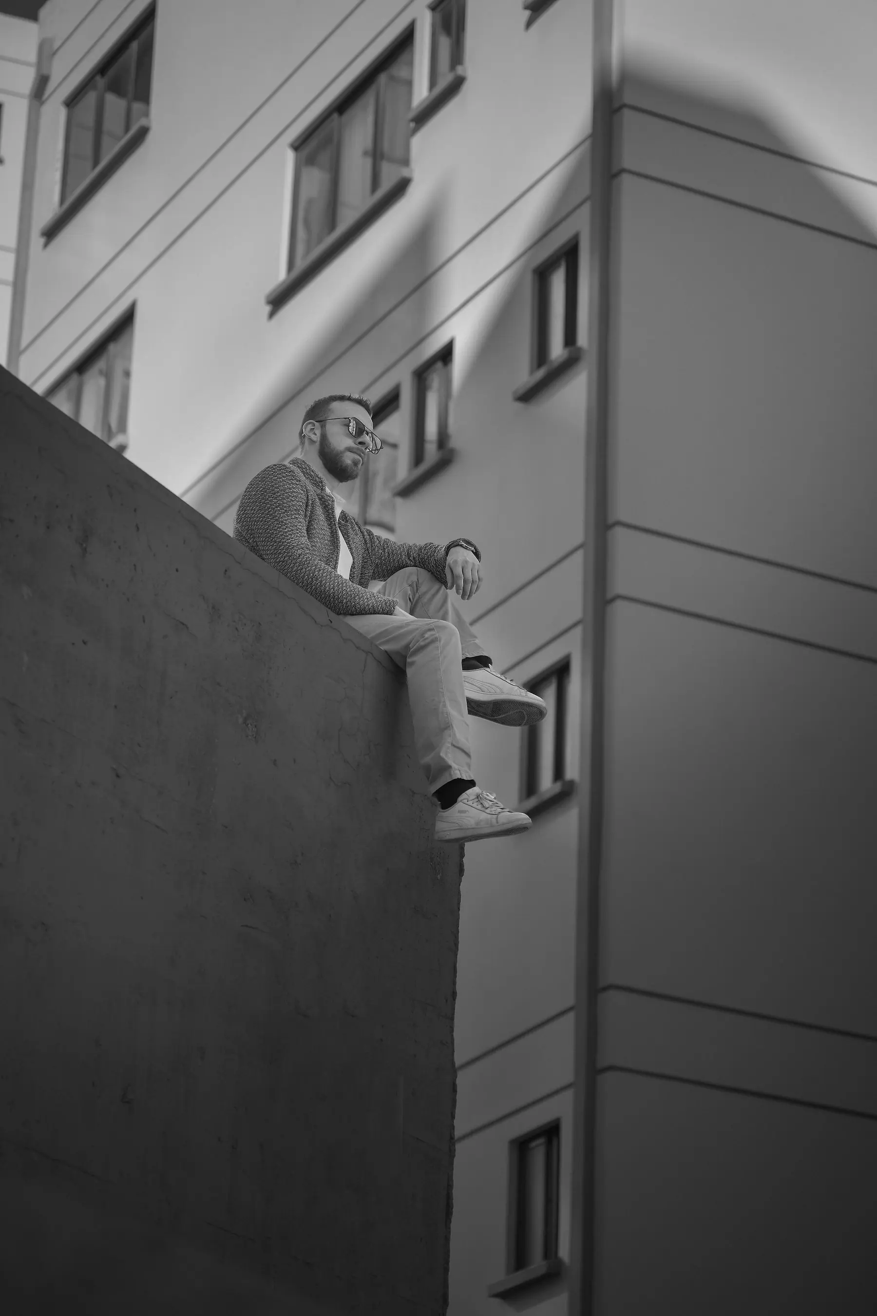 Fotografía en blanco y negro de Elex Asbun sentado en el borde de un muro alto, con la fachada de un edificio moderno detrás.