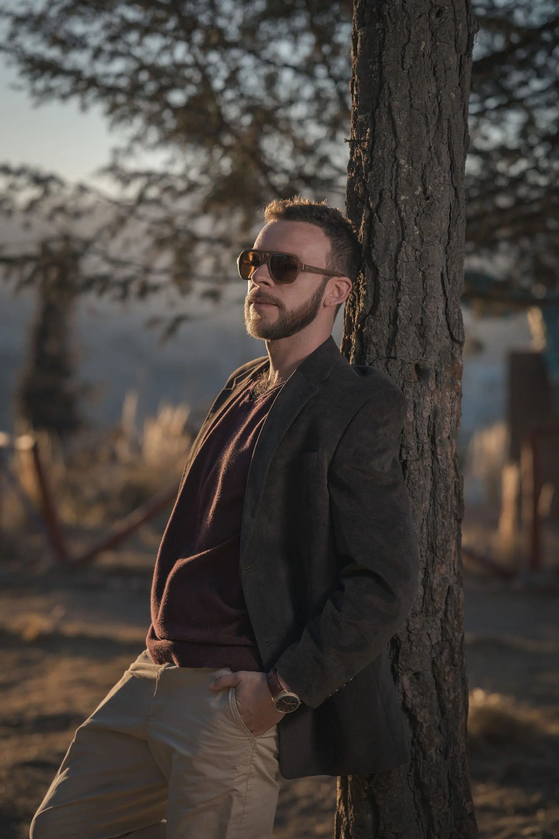 Elex Asbun con gafas de sol, blazer marrón y jersey burdeos, apoyado en el tronco de un pino a contraluz del atardecer.